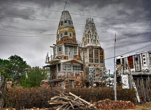 A unique, weathered house with two tall towers, surrounded by overgrown vegetation and scattered debris under a cloudy sky.