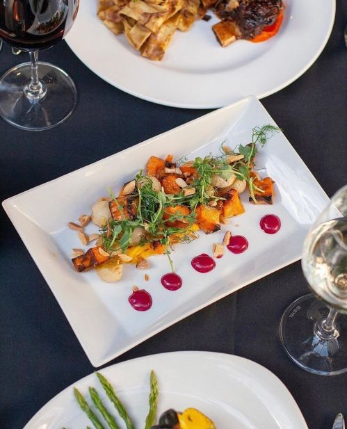 A gourmet dish featuring roasted vegetables, microgreens, and sauce, served on a white plate with wine glasses in the background.