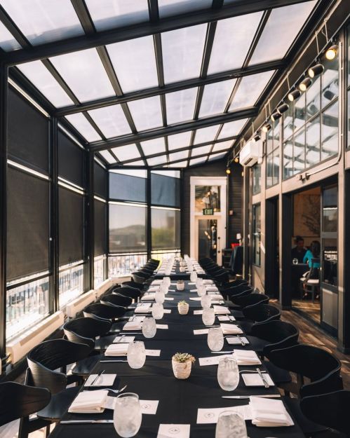 A long dining table set with glasses and napkins in a bright, glass-enclosed restaurant space.