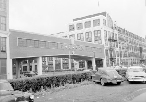 Historic Packard factory entrance with vintage cars parked outside, surrounded by industrial buildings. Black and white photo.