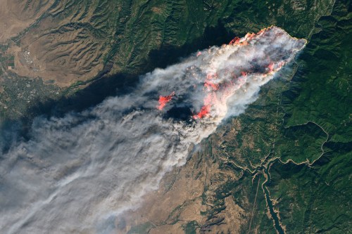 Aerial view of a large wildfire, with smoke and flames spreading across a mountainous landscape.