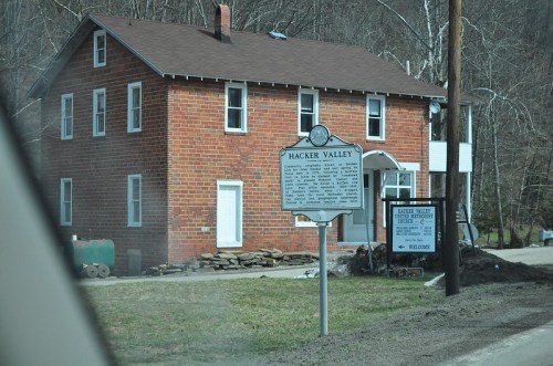 A brick house in Hacker Valley, with a historical marker and a welcome sign in front, surrounded by trees.
