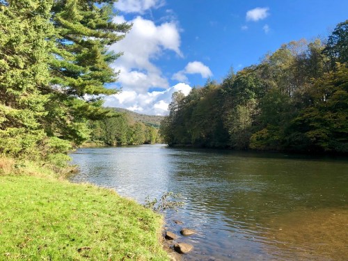 A serene river flows through a lush landscape, surrounded by trees and under a partly cloudy blue sky.