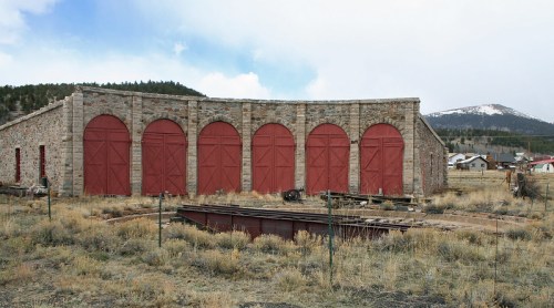 Historic stone building with red doors, surrounded by dry grass and mountains in the background.