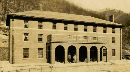 Historic brick building with arched entrance, two people standing outside, surrounded by hills. Black and white photo.
