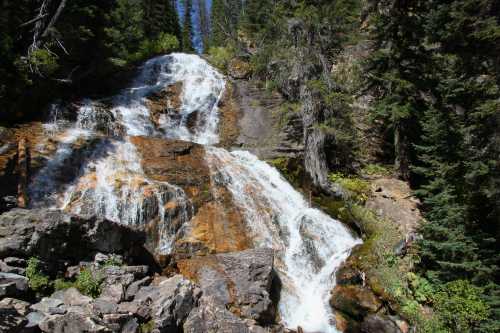 A cascading waterfall flows over rocky terrain, surrounded by lush green trees and a clear blue sky.