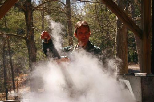 A man in a forest holds a container, surrounded by steam and mist from a nearby boiling pot.