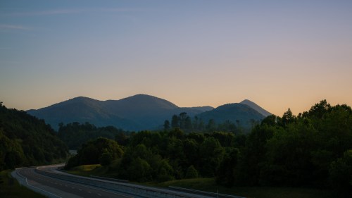 A serene landscape featuring rolling mountains at dusk, with a highway winding through lush greenery in the foreground.