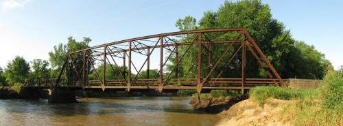 A rusted metal bridge spans a river, surrounded by lush green trees and a sandy bank under a clear blue sky.