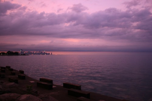 A serene lakeside view at dusk, with a city skyline silhouetted against a purple and gray sky.