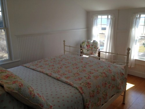 Bright bedroom with a floral quilt, white walls, and large windows letting in natural light. A floral chair is in the corner.