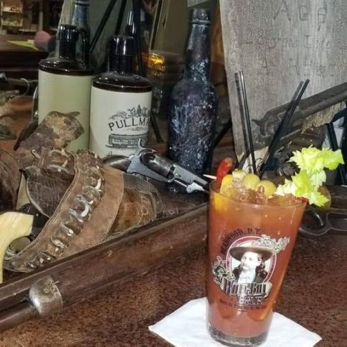 A colorful cocktail garnished with fruit sits on a rustic bar, surrounded by vintage bottles and western-themed decor.