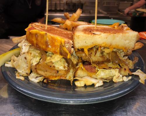 A close-up of a grilled sandwich filled with cabbage, cheese, and onions, served with fries and a pickle on a plate.