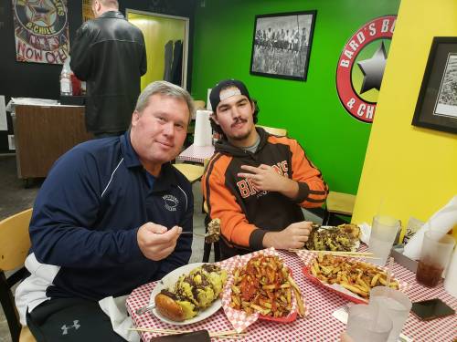 Two men sit at a table in a colorful restaurant, enjoying large plates of food and fries.