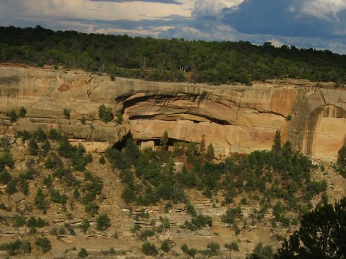 A rocky cliff with layered formations and sparse vegetation under a cloudy sky.