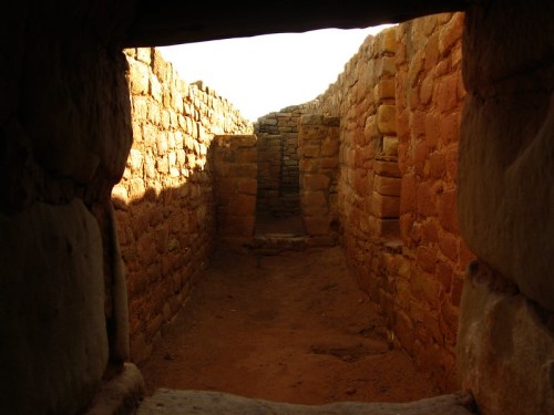 View through a stone doorway into a narrow, sunlit corridor of ancient ruins with textured walls.