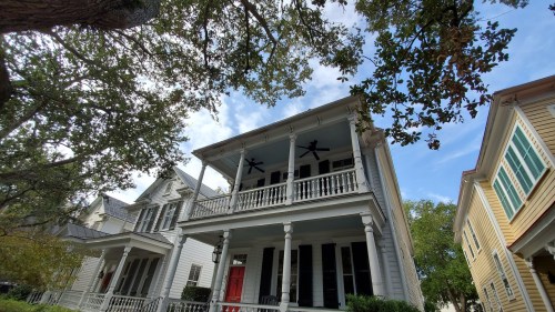 A two-story house with a red door, large porch, and ceiling fans, surrounded by trees and other historic homes.