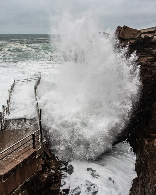 A powerful wave crashes against a rocky shore, splashing over a pier under a cloudy sky.