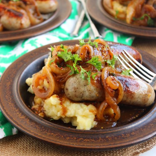 A plate of sausages topped with caramelized onions, served over creamy mashed potatoes, garnished with parsley.