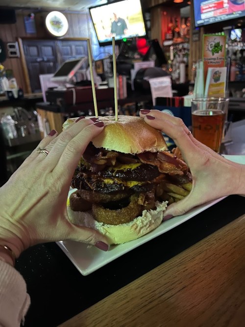 A person holds a towering burger with bacon, cheese, and onion rings, served with fries in a bar setting.