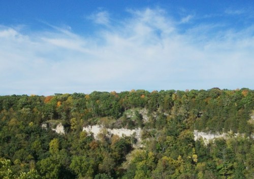 A scenic view of a forested cliff under a blue sky with scattered clouds, showcasing autumn foliage.