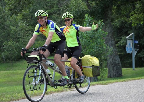 A couple rides a tandem bicycle, both wearing bright yellow cycling gear, smiling and enjoying a sunny day outdoors.