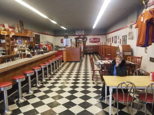 A retro diner with checkered floors, red stools, a jukebox, and a woman sitting at a table.