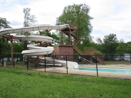 A water slide next to a pool, surrounded by a fence and greenery, with a building and stairs in the background.