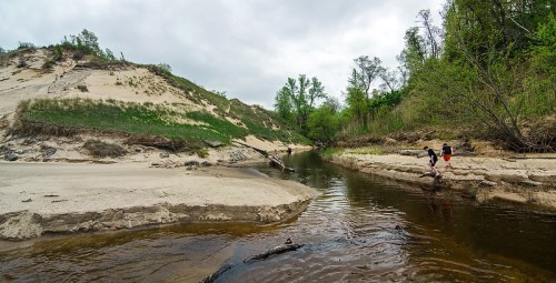 A serene river flows between sandy banks, surrounded by lush greenery and overcast skies. Two figures walk along the shore.