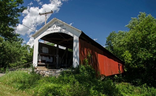 A historic covered bridge with a red exterior, surrounded by greenery and under a blue sky with clouds.