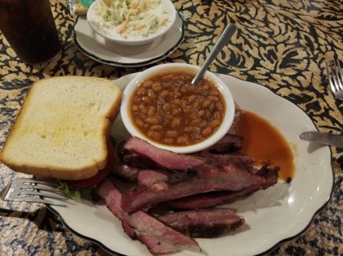 A plate with sliced brisket, baked beans in a bowl, a slice of bread, and a side of coleslaw on a patterned tablecloth.