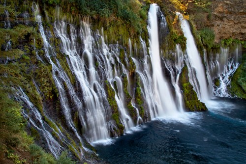 A serene waterfall cascades over moss-covered rocks into a deep blue pool, surrounded by lush greenery.
