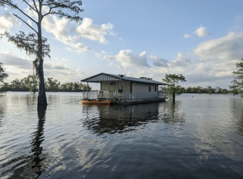 A houseboat on calm water surrounded by trees, with a cloudy sky reflecting in the water.