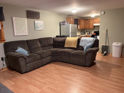 Cozy living room featuring a brown sectional sofa, light blue pillows, and a kitchen area in the background.