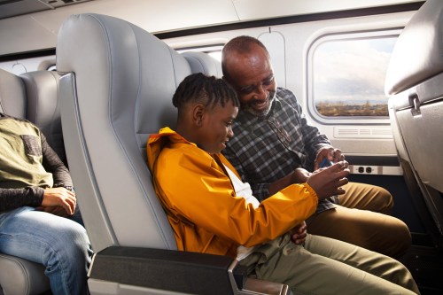 A man and a child share a moment on a train, looking at a device together, while another passenger sits nearby.