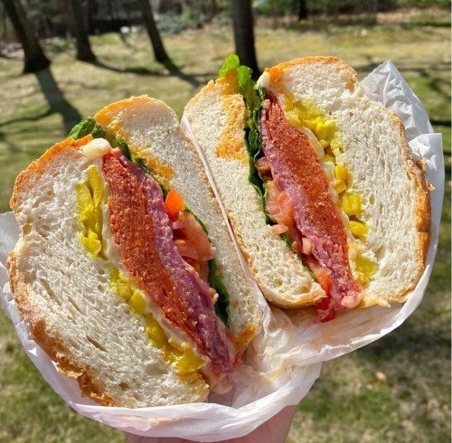 A hand holding a freshly cut sandwich filled with lettuce, tomato, pickles, and layers of meat on soft bread.