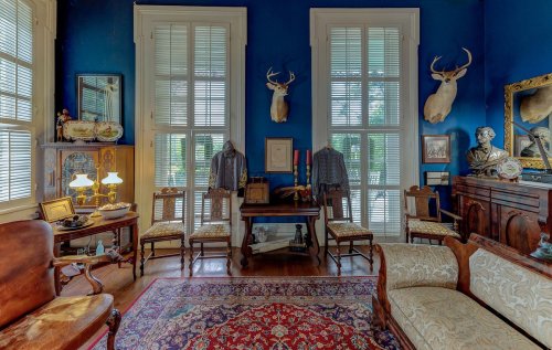 A vintage room with blue walls, antique furniture, deer heads on the wall, and historical artifacts displayed.