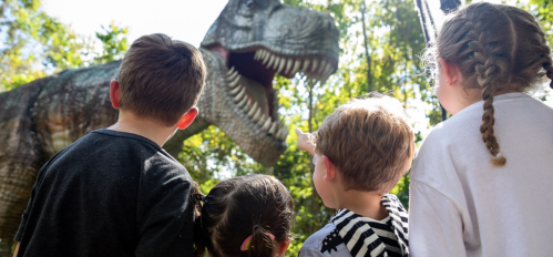Four children gaze in awe at a large dinosaur statue in a lush, green outdoor setting.