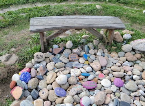 A wooden bench beside a colorful collection of painted rocks in a grassy area.