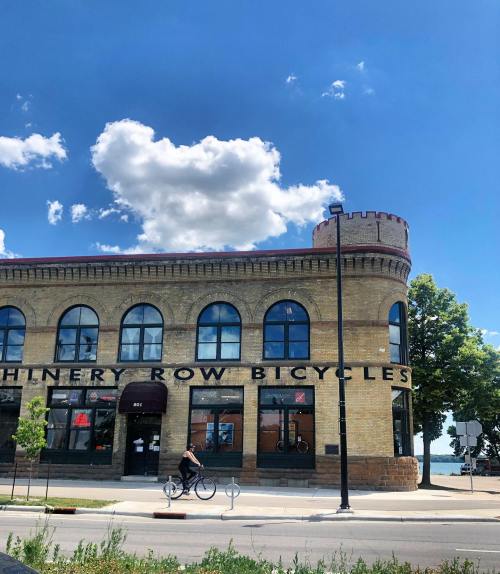 A brick building with arched windows, labeled "Machinery Row Bicycles," under a blue sky with fluffy clouds.