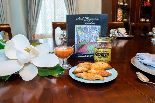 A table setting featuring a book, a jar of salsa, a cocktail, and savory pastries, with a large white flower nearby.