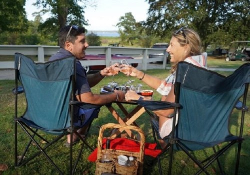 A couple toasting with glasses while sitting in camping chairs, surrounded by a picnic setup in a scenic outdoor setting.