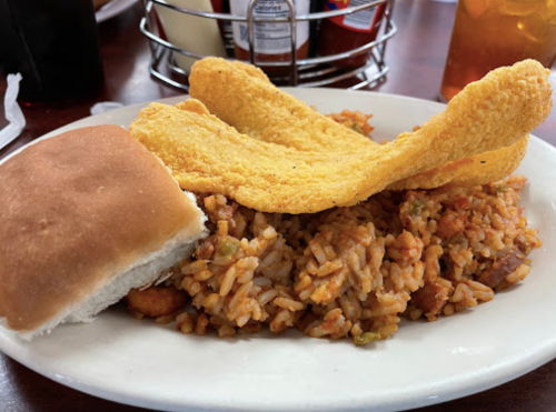 A plate of rice with spices, topped with two crispy fish fillets and a soft bread roll on the side.