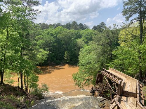 A wooden waterwheel beside a muddy river, surrounded by lush green trees and a bright blue sky with clouds.