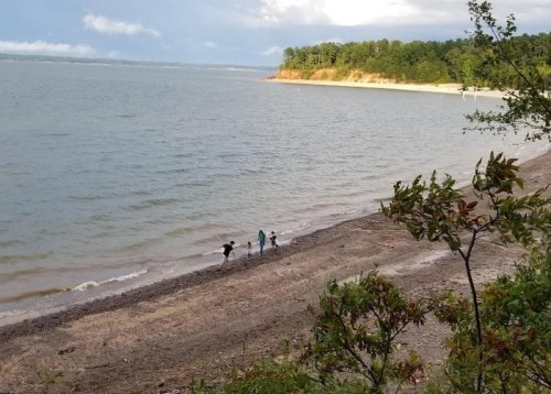 Three people walking along a sandy beach by a calm lake, surrounded by trees and a cloudy sky.