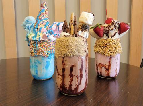 Three colorful milkshakes topped with whipped cream, sprinkles, cookies, and fruit, served in tall glasses on a table.