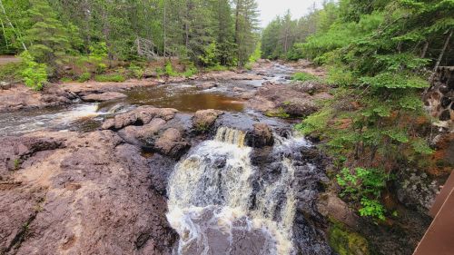 A serene river scene with small waterfalls surrounded by lush green trees and rocky terrain.