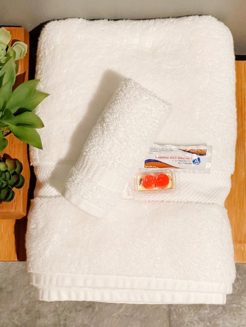 A neatly arranged set of white towels with a small container of ointment and a plant in the background.