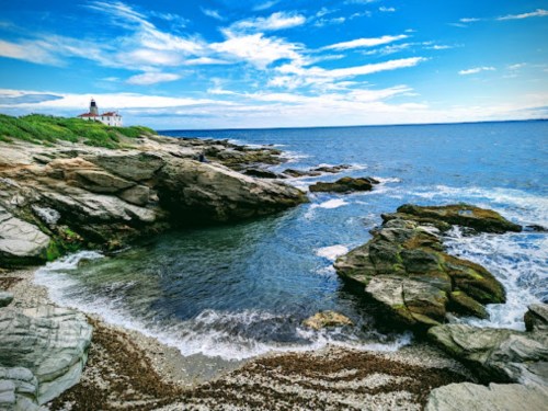 A rocky coastline with clear water, featuring a lighthouse in the distance under a bright blue sky.