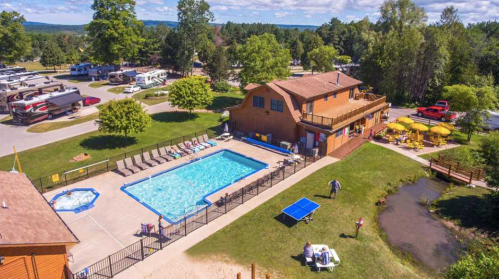 Aerial view of a pool area with lounge chairs, surrounded by trees and RVs, featuring a building and picnic tables nearby.